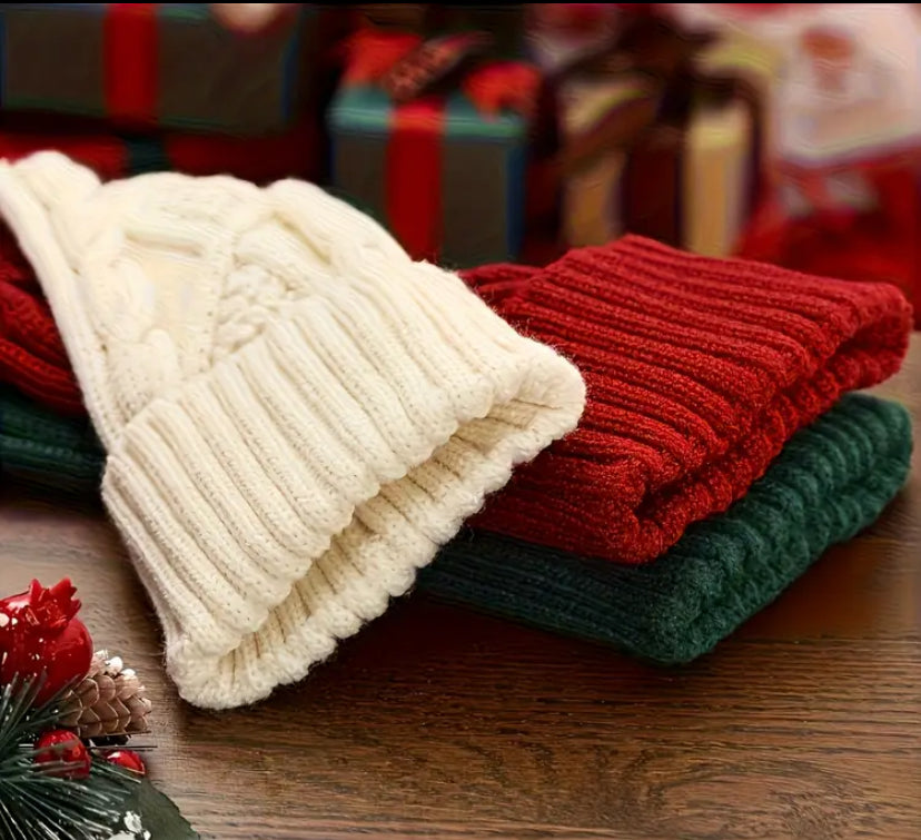 Stack of red, green, and white knitted Christmas stockings on wooden table with festive decorations and gift boxes in the background.